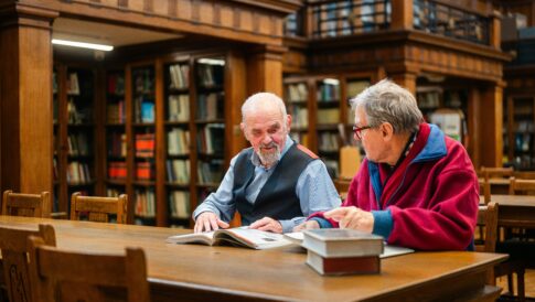 Zdjęcie przedstawia dwóch mężczyzn w starszym wieku, którzy siedzą obok siebie przy stole w czytelni bibliotecznej. Przed nimi leżą książki - jedna otwarta i kilka zamkniętych.