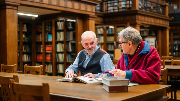 Zdjęcie przedstawia dwóch mężczyzn w starszym wieku, którzy siedzą obok siebie przy stole w czytelni bibliotecznej. Przed nimi leżą książki - jedna otwarta i kilka zamkniętych.