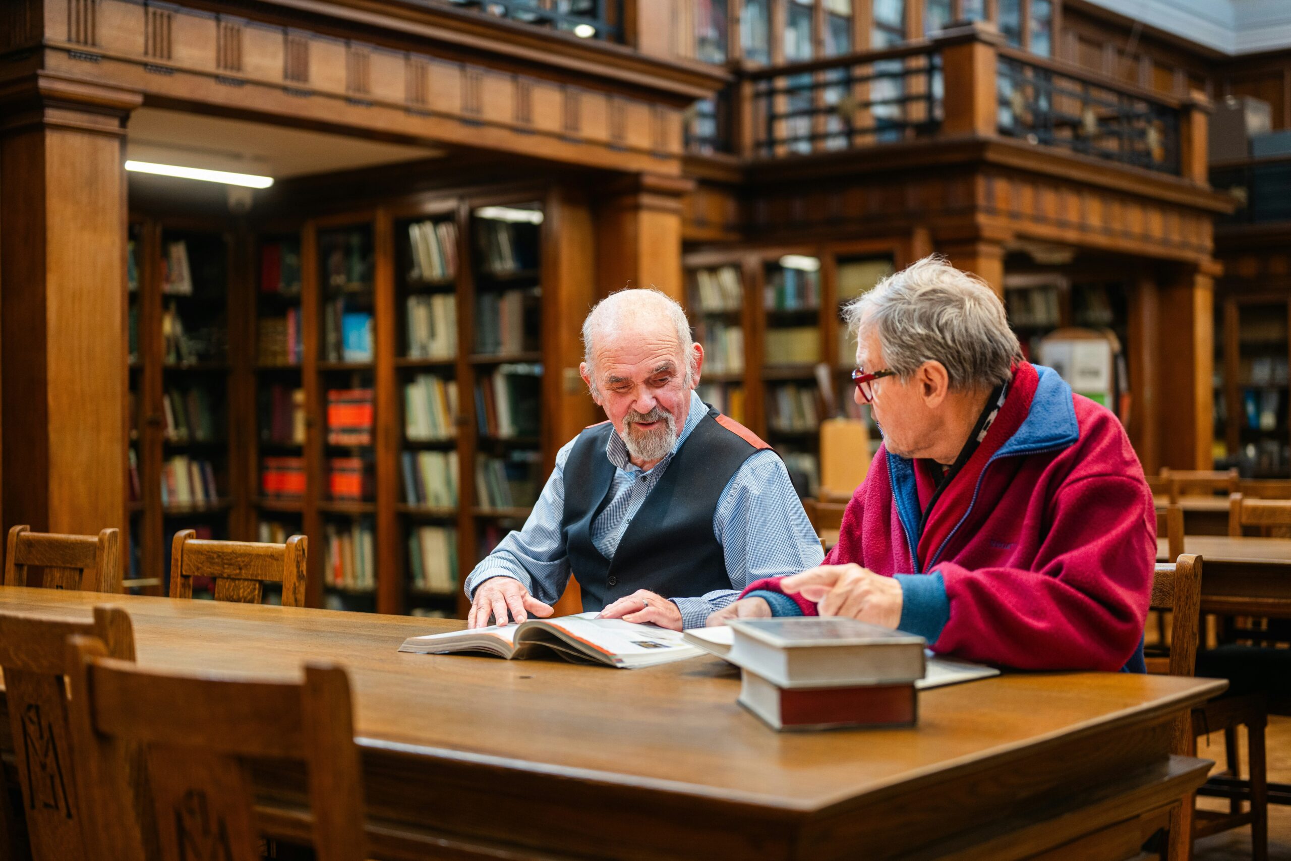 Zdjęcie przedstawia dwóch mężczyzn w starszym wieku, którzy siedzą obok siebie przy stole w czytelni bibliotecznej. Przed nimi leżą książki - jedna otwarta i kilka zamkniętych.