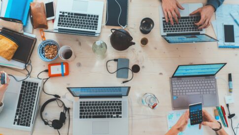 Grupa ludzi pracuje przy jasnym drewnianym stole z otwartymi laptopami, notatnikami, przekąskami, napojami, słuchawkami, smartfonem i papierami. Widoczne są ręce piszące na klawiaturze, piszące i korzystające z telefonu, co pokazuje ruchliwe, oparte na współpracy środowisko pracy.