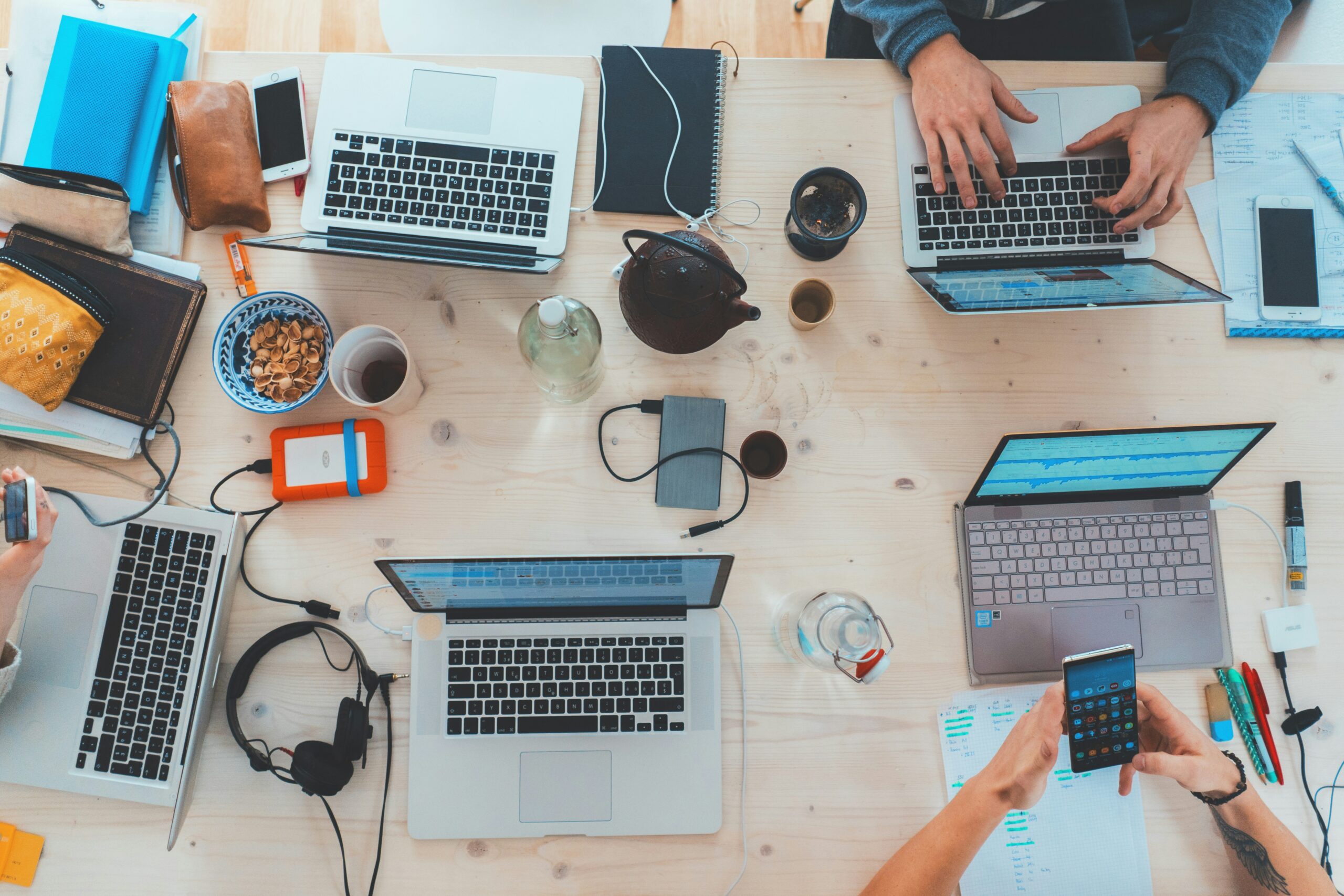 Grupa ludzi pracuje przy jasnym drewnianym stole z otwartymi laptopami, notatnikami, przekąskami, napojami, słuchawkami, smartfonem i papierami. Widoczne są ręce piszące na klawiaturze, piszące i korzystające z telefonu, co pokazuje ruchliwe, oparte na współpracy środowisko pracy.