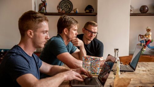 Zdjęcie przedstawia trzech mężczyzn siedzących przy stole. Mężczyżni siedzą przy komputerach - jeden mężczyzna siedzi sam przy komputerze, a obok dwóch mężczyzn patrzy na ekran drugiego laptopa.